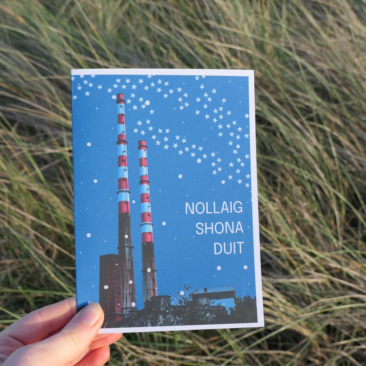 Hand holding a blue greeting card for Christmas with text and an image of the Poolbeg Stacks in Dublin against a natural background