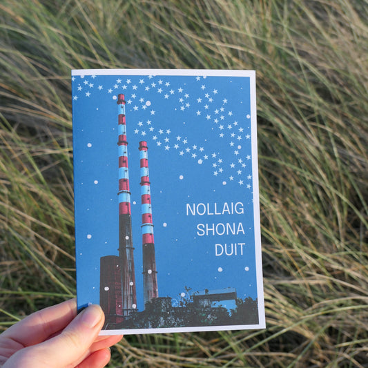 Hand holding a blue greeting card for Christmas with text and an image of the Poolbeg Stacks in Dublin against a natural background