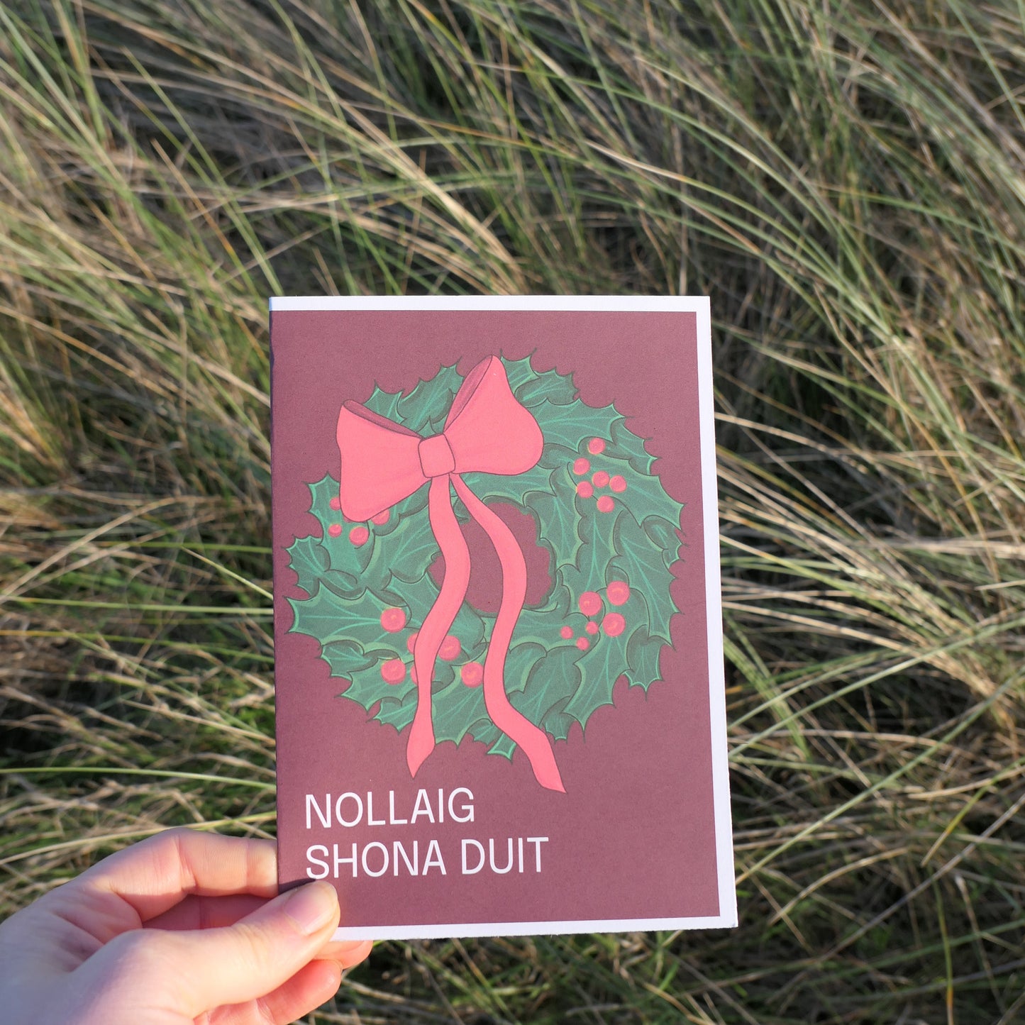 Hand holding a Christmas card with a wreath and bow design against a grassy background